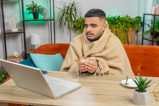 Sick ill Indian man freelancer working on laptop wrapped in plaid sit shivering from cold drinking hot coffee in unheated home office without heating due debt. Unhealthy Hindu guy try to warming up