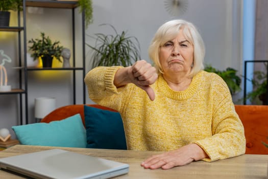 Dislike. Upset unhappy senior woman showing thumbs down sign gesture, expressing discontent, disapproval dissatisfied bad work at home apartment indoors. Displeased elderly grandmother sits at table