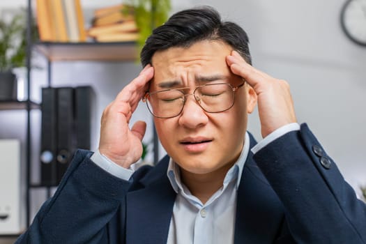 Close-up face of tired ill Asian businessman suffering from headache problem tension and migraine, stress at office desk. Mature freelancer broker worker man works on laptop notebook. Overworking