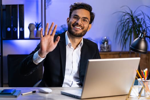 Hello. Indian business man working on laptop computer smiling friendly at camera and waving hands gesturing hi, greeting or goodbye, welcoming with hospitable expression at home office workplace desk