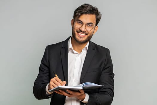 Thoughtful journalist Indian businessman making notes, writing down thoughts with pen into notepad notebook diary, to do list, good idea. Male Arabian man guy lawyer isolated alone on gray background