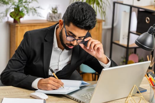 Focused serious Indian businessman freelancer working at modern home office workplace desk writing notes ideas in notebook. Male Arabian in formal suit preparing planning presentation startup project