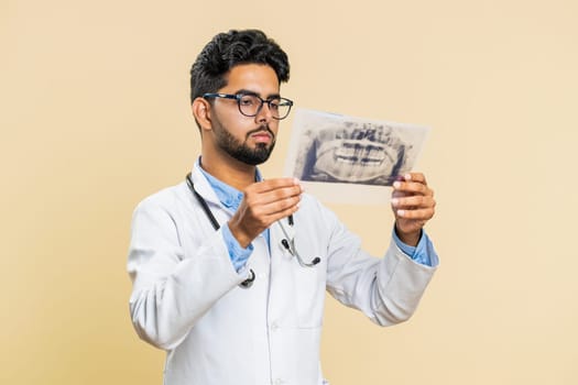 Indian young doctor orthodontist man examines a panoramic x-ray picture of the jaw teeth. 3D model of the patient's mouth, MRI scan. Dentistry, oral care. Arabian stomatology guy on beige background