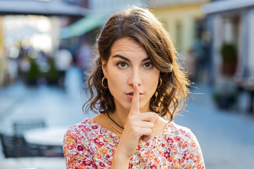 Shh be quiet please. Caucasian young woman presses index finger to lips makes silence hush gesture sign do not tells gossip secret outdoors. Strict girl standing in urban city street. Town lifestyles