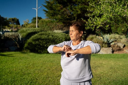 Determined senior woman raises her arms while exercising at park on sunny day, embracing energy of outdoors as she focuses on her fitness and strength