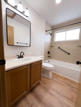 Broomfield, Colorado, USA-January 25, 2025-A modern bathroom featuring a white bathtub with beige tiled walls, a vanity with a white sink, and black fixtures. The bathroom includes a large mirror, a three-light vanity fixture, and a window allowing natural light. The wooden cabinets and light wood flooring complement the neutral color scheme. Black towel bars, a shower rod, and grab bars add a stylish contrast.