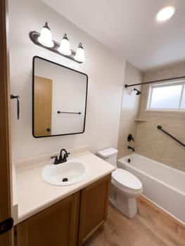 Broomfield, Colorado, USA-January 25, 2025-A modern bathroom featuring a white bathtub with beige tiled walls, a vanity with a white sink, and black fixtures. The bathroom includes a large mirror, a three-light vanity fixture, and a window allowing natural light. The wooden cabinets and light wood flooring complement the neutral color scheme. Black towel bars, a shower rod, and grab bars add a stylish contrast.