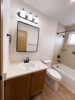 Broomfield, Colorado, USA-January 25, 2025-A modern bathroom featuring a white bathtub with beige tiled walls, a vanity with a white sink, and black fixtures. The bathroom includes a large mirror, a three-light vanity fixture, and a window allowing natural light. The wooden cabinets and light wood flooring complement the neutral color scheme. Black towel bars, a shower rod, and grab bars add a stylish contrast.