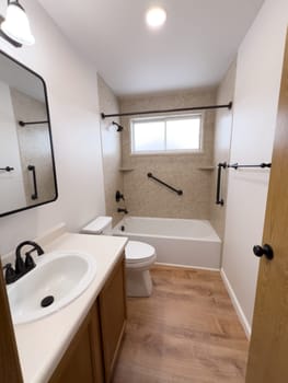 Broomfield, Colorado, USA-January 25, 2025-A modern bathroom featuring a white bathtub with beige tiled walls, a vanity with a white sink, and black fixtures. The bathroom includes a large mirror, a three-light vanity fixture, and a window allowing natural light. The wooden cabinets and light wood flooring complement the neutral color scheme. Black towel bars, a shower rod, and grab bars add a stylish contrast.