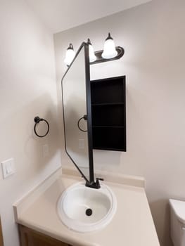 Broomfield, Colorado, USA-January 25, 2025-A stylish bathroom vanity featuring a large rectangular mirror with a black frame, a white sink with a black faucet, and a beige countertop. The vanity is complemented by a three-light fixture above the mirror, providing ample lighting. A black towel bar is mounted on the wall, enhancing the modern design. The bathroom has a clean, minimalist look with neutral tones and functional elements.
