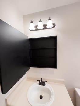 Broomfield, Colorado, USA-January 25, 2025-A stylish bathroom vanity featuring a large rectangular mirror with a black frame, a white sink with a black faucet, and a beige countertop. The vanity is complemented by a three-light fixture above the mirror, providing ample lighting. A black towel bar is mounted on the wall, enhancing the modern design. The bathroom has a clean, minimalist look with neutral tones and functional elements.