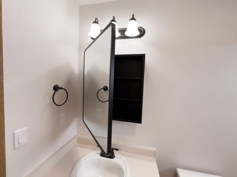 Broomfield, Colorado, USA-January 25, 2025-A stylish bathroom vanity featuring a large rectangular mirror with a black frame, a white sink with a black faucet, and a beige countertop. The vanity is complemented by a three-light fixture above the mirror, providing ample lighting. A black towel bar is mounted on the wall, enhancing the modern design. The bathroom has a clean, minimalist look with neutral tones and functional elements.