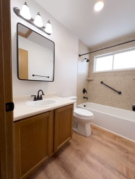 Broomfield, Colorado, USA-January 25, 2025-A modern bathroom featuring a white bathtub with beige tiled walls, a vanity with a white sink, and black fixtures. The bathroom includes a large mirror, a three-light vanity fixture, and a window allowing natural light. The wooden cabinets and light wood flooring complement the neutral color scheme. Black towel bars, a shower rod, and grab bars add a stylish contrast.