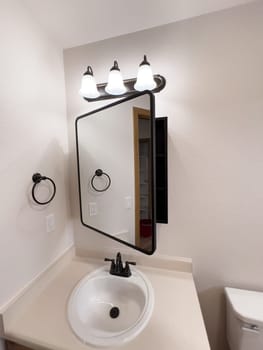 Broomfield, Colorado, USA-January 25, 2025-A stylish bathroom vanity featuring a large rectangular mirror with a black frame, a white sink with a black faucet, and a beige countertop. The vanity is complemented by a three-light fixture above the mirror, providing ample lighting. A black towel bar is mounted on the wall, enhancing the modern design. The bathroom has a clean, minimalist look with neutral tones and functional elements.