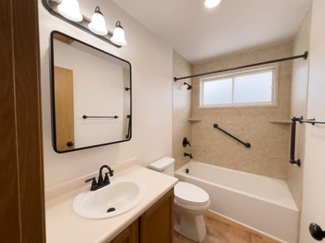 Broomfield, Colorado, USA-January 25, 2025-A modern bathroom featuring a white bathtub with beige tiled walls, a vanity with a white sink, and black fixtures. The bathroom includes a large mirror, a three-light vanity fixture, and a window allowing natural light. The wooden cabinets and light wood flooring complement the neutral color scheme. Black towel bars, a shower rod, and grab bars add a stylish contrast.