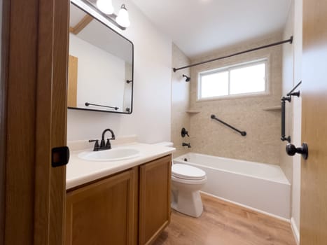 Broomfield, Colorado, USA-January 25, 2025-A modern bathroom featuring a white bathtub with beige tiled walls, a vanity with a white sink, and black fixtures. The bathroom includes a large mirror, a three-light vanity fixture, and a window allowing natural light. The wooden cabinets and light wood flooring complement the neutral color scheme. Black towel bars, a shower rod, and grab bars add a stylish contrast.