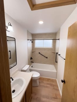 Broomfield, Colorado, USA-January 25, 2025-A modern bathroom featuring a white bathtub with beige tiled walls, a vanity with a white sink, and black fixtures. The bathroom includes a large mirror, a three-light vanity fixture, and a window allowing natural light. The wooden cabinets and light wood flooring complement the neutral color scheme. Black towel bars, a shower rod, and grab bars add a stylish contrast.