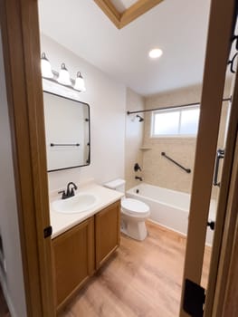 Broomfield, Colorado, USA-January 25, 2025-A modern bathroom featuring a white bathtub with beige tiled walls, a vanity with a white sink, and black fixtures. The bathroom includes a large mirror, a three-light vanity fixture, and a window allowing natural light. The wooden cabinets and light wood flooring complement the neutral color scheme. Black towel bars, a shower rod, and grab bars add a stylish contrast.