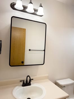 Broomfield, Colorado, USA-January 25, 2025-A stylish bathroom vanity featuring a large rectangular mirror with a black frame, a white sink with a black faucet, and a beige countertop. The vanity is complemented by a three-light fixture above the mirror, providing ample lighting. A black towel bar is mounted on the wall, enhancing the modern design. The bathroom has a clean, minimalist look with neutral tones and functional elements.