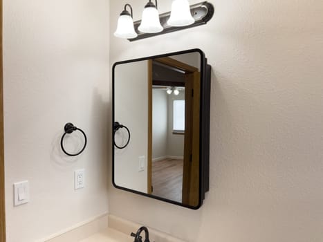 Broomfield, Colorado, USA-January 25, 2025-A stylish bathroom vanity featuring a large rectangular mirror with a black frame, a white sink with a black faucet, and a beige countertop. The vanity is complemented by a three-light fixture above the mirror, providing ample lighting. A black towel bar is mounted on the wall, enhancing the modern design. The bathroom has a clean, minimalist look with neutral tones and functional elements.