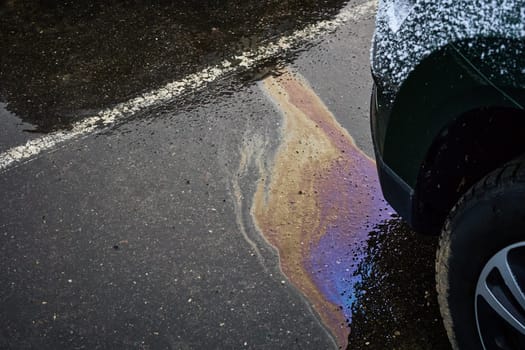 Colorful oil leak forming an iridescent sheen on the damp asphalt near a vehicle, drawing attention to pollution concerns.