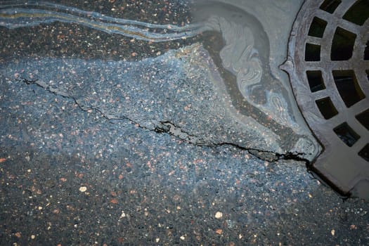 Environmental pollution creating a colorful sheen near a storm drain on a cracked asphalt road