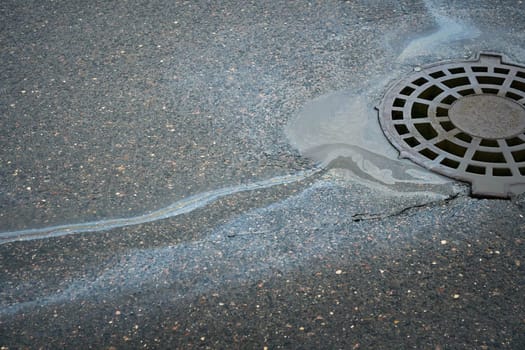 Gasoline or oil spill creating rainbow sheen near storm drain on asphalt road.