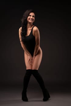 front view of sexy female in black bodysuit standing in studio on dark background and smile at camera