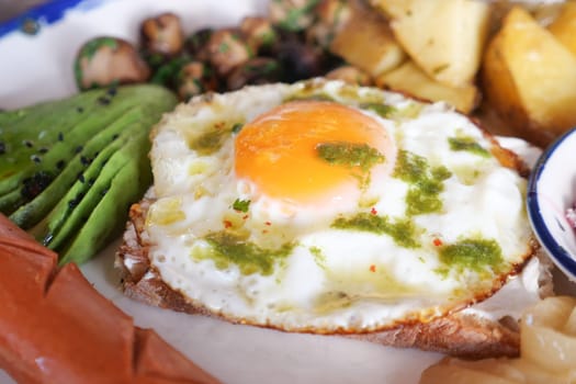 A colorful breakfast plate featuring a fried egg, avocado, and sausages.