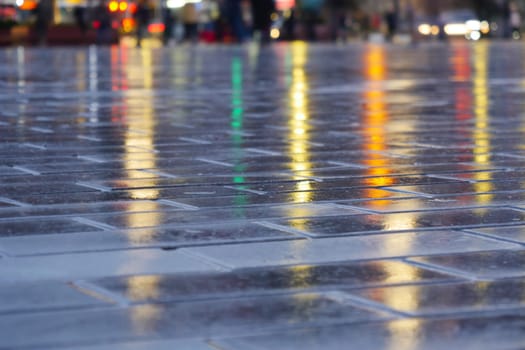 Colorful lights reflect on a rain-soaked pavement during evening hours.