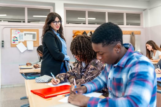 Multi ethnic high school students focused on writing exams in a classroom, with a teacher supervising and providing guidance