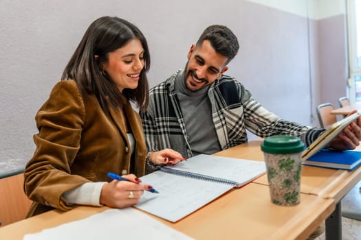 Two happy university students are studying together in a classroom, taking notes and using a tablet, enjoying the learning process