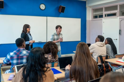 Male high school student stands and gives a presentation to his diverse classmates with his teacher standing beside him in their modern classroom