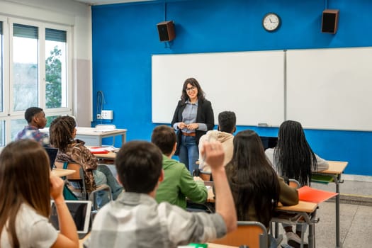 Multiethnic students raising hands during a lesson held by female teacher in high school classroom