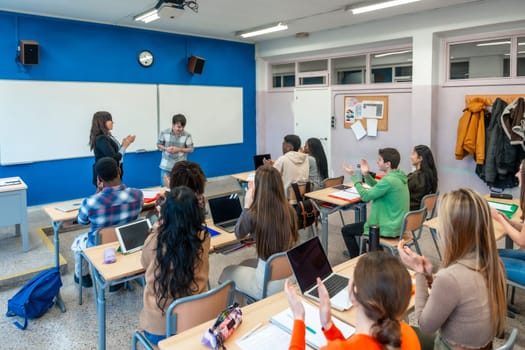 Multi ethnic university students clapping in classroom after a presentation of a shy student, with teacher congratulating him