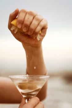 Cropped hand squeezing lemon juice to a glass of see-through yellow cocktail. Lots of lemons. Moody still life. Refreshing drink. High quality photo