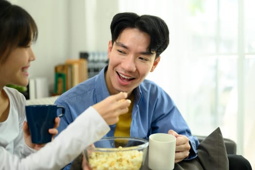 Happy young couple sitting on the couch, sharing a bowl of popcorn while watching movie.