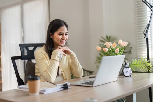 A smiling young woman engaging with her laptop in a sunny home office, surrounded by flowers and greenery, radiating positivity and productivity.