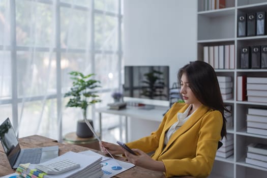 Young businesswoman working in modern office with laptop, papers, and smartphone on a bright day.