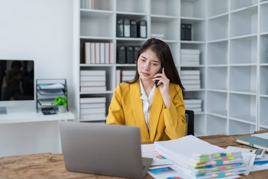 Focused businesswoman in a yellow blazer engaging in a phone conversation while working on a laptop in a modern office.