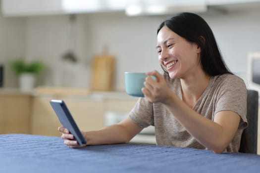 Happy asian woman reading ebook drinking coffee in the kitchen at home