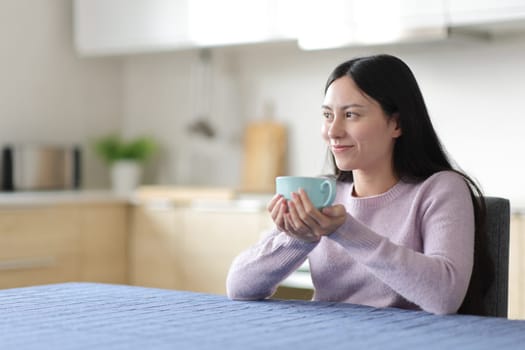 Relaxed asian woman drinking and contemplating in the kitchen at home