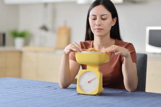 Front view portrait of a serious asian woman weighing food using scale in a kitchen at home