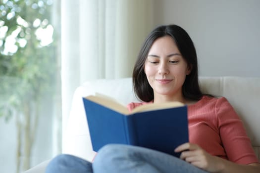 Satisfied asian woman reading a paper book sitting on a couch at home