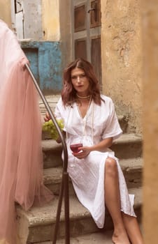 young woman eating grapes and drinking red wine. she is wearing a summer white dress. sitting on the steps of an old building. holding a bunch of grapes and a glass of wine and looking at the camera