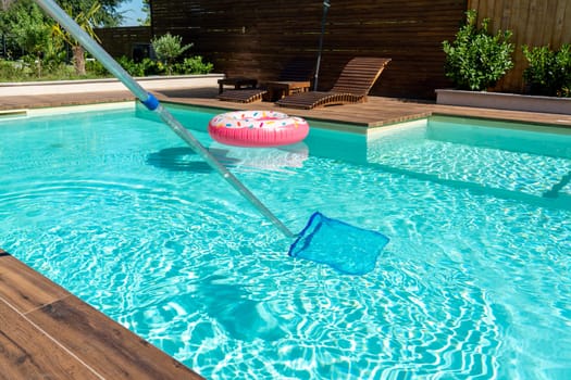 Skimmer net cleaning turquoise swimming pool water with wooden deck and colorful inflatable ring