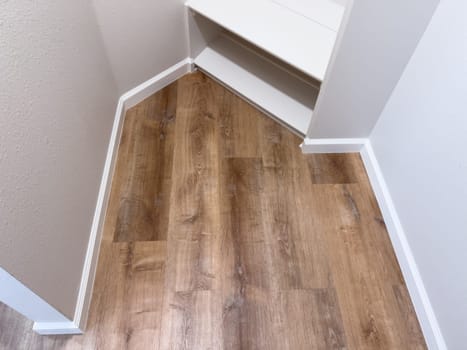 A small closet corner featuring white built-in shelves and wood-look vinyl flooring. The walls are painted white, and the baseboards provide a clean, modern finish. The space is well-lit, showcasing a simple and functional storage solution.