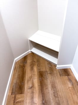 A small closet corner featuring white built-in shelves and wood-look vinyl flooring. The walls are painted white, and the baseboards provide a clean, modern finish. The space is well-lit, showcasing a simple and functional storage solution.
