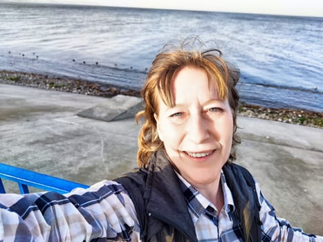 Middle-aged woman captures a joyful selfie with the beautiful water view behind her during a summer excursion.
