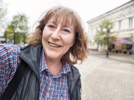 A middle-aged blonde woman enjoys a fun moment taking a selfie while exploring a quaint city street.